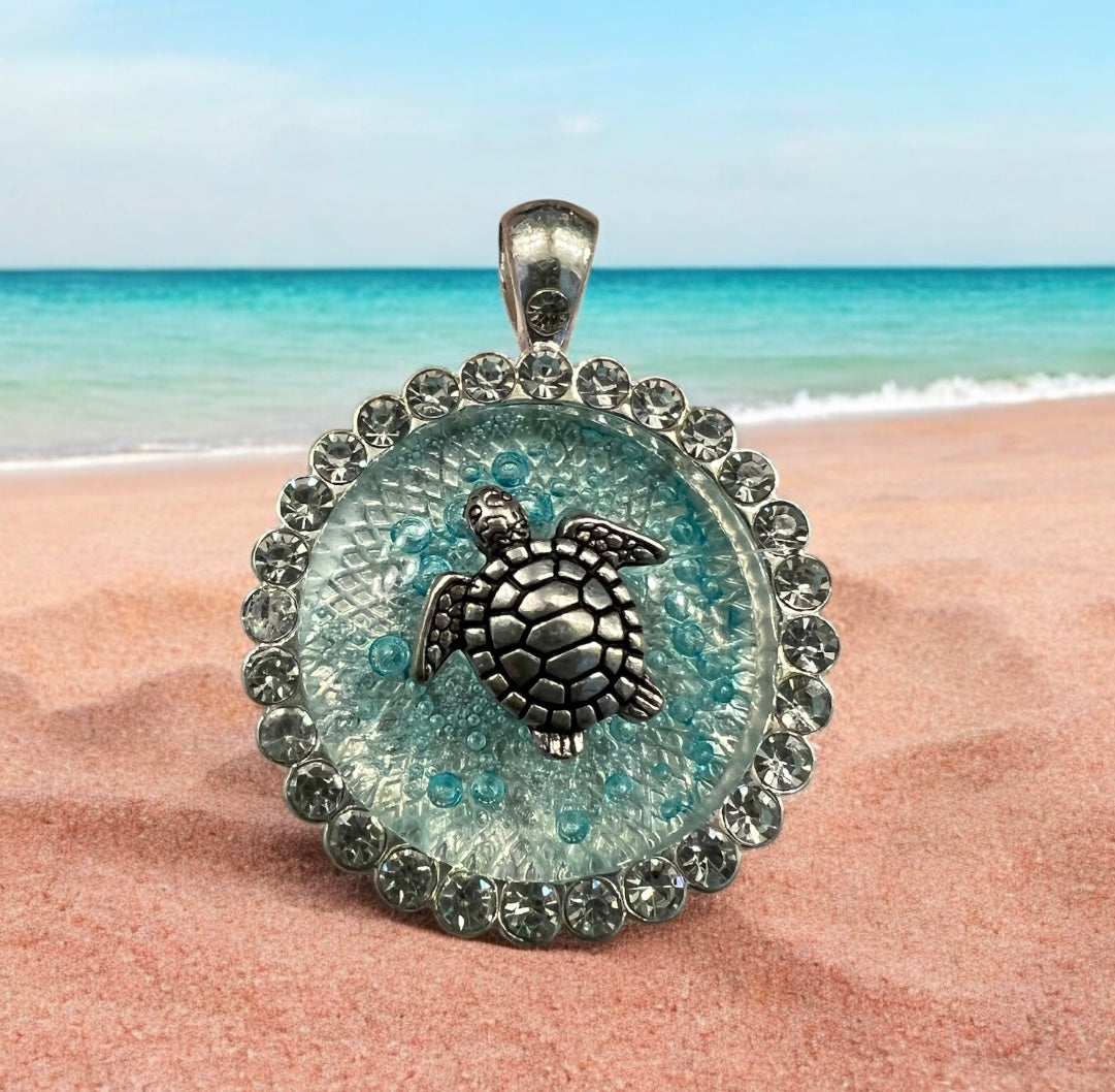 Stunningly clear sea-blue turtle necklace on a sandy beach with the ocean in the background.